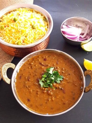 Dal Makhani garnished with cilantro leaves served in copper bowl. A bowl of lemon rice and a bowl of cut red onion and lemons in the background.