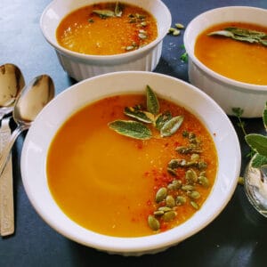 A soup bowl full of creamy golden butternut squash soup. It is garnished with sage and pumpkin seeds.