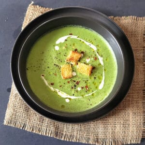 A soup bowl filled with creamy asparagus broccoli soup garnished with olive oil, light cream and croutons.