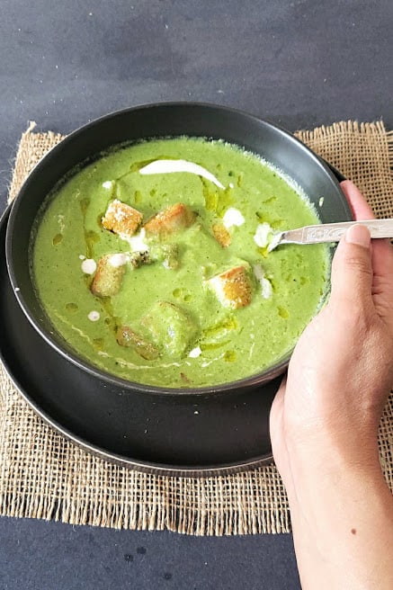 A hand holding black soup bowl full of seasoned creamy soup and the spoon dunked in the soup.