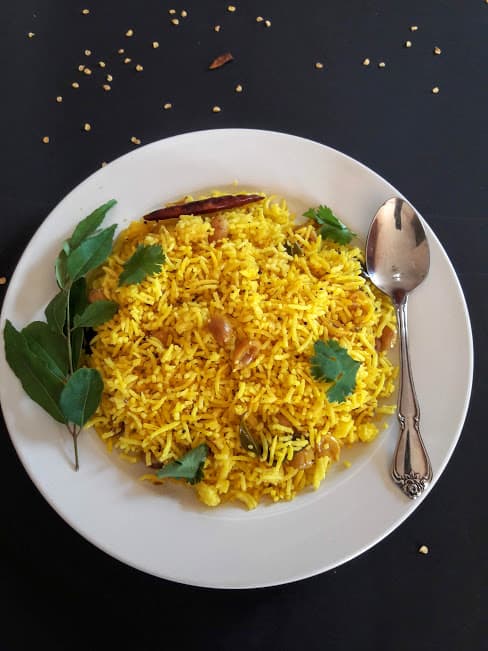 Golden yellow rice served on dinner plate along with curry leaves strand and a spoon.