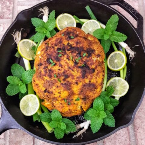 Whole Roasted Cauliflower served on black cast iron skillet with mint, lemon wedges and green onion garnish.