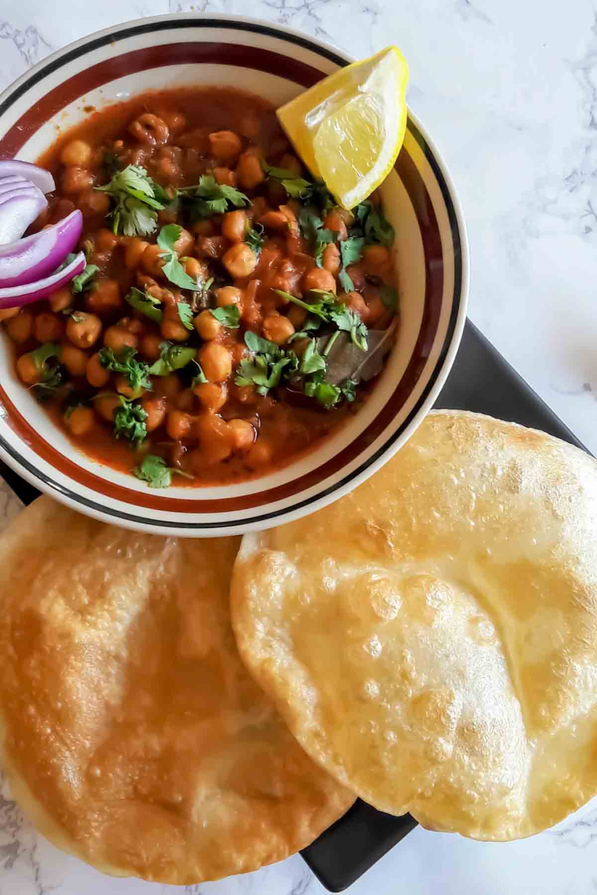 Chole curry and Bhature on black plate with lemon wedges and onion and chili.