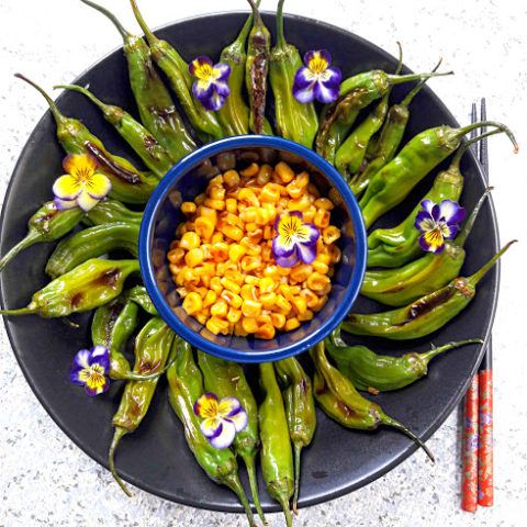 Grilled Shishito peppers served on black platter with grilled corn in the center with viola flowers spirinkle is recipe from profusioncurry