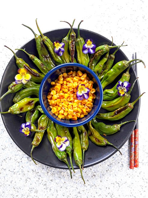 Grilled Shishito peppers served on black platter with grilled corn in the center with viola flowers spirinkle is recipe from profusioncurry