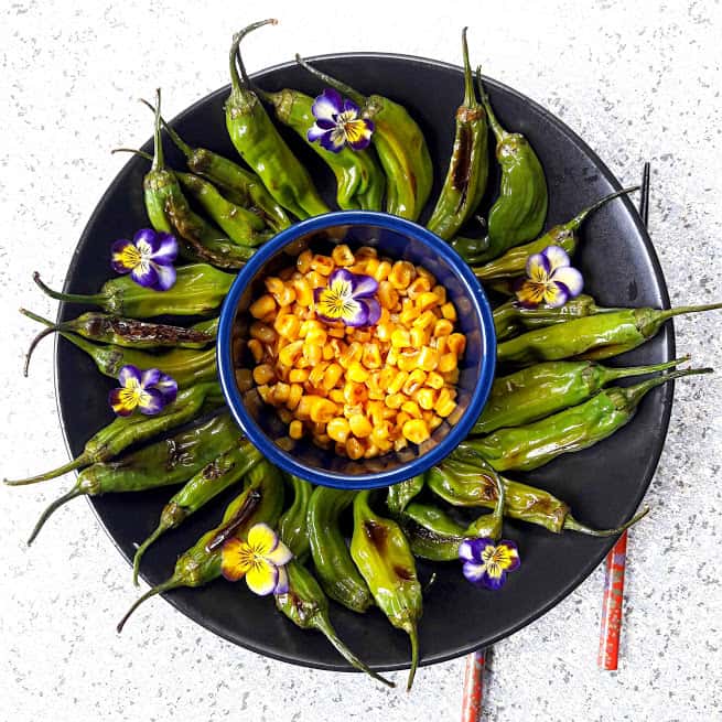 Grilled Shishito peppers served on black platter with grilled corn in the center with viola flowers spirinkle is recipe from profusioncurry 