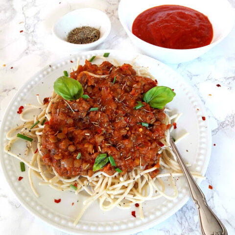 Close up photo of lentil bolognese sauce served over gluten free spaghetti. The dinner is served in white dinner plate with fresh basil leaves and red chili flakes garnish.