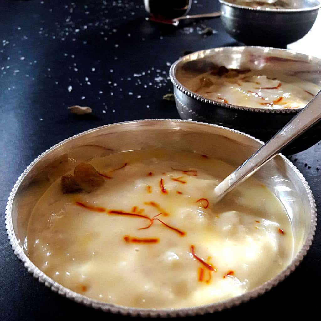 Silver bowl and spoon containing Indian dessert of coconut rice kheer garnished with saffron and raisins. 