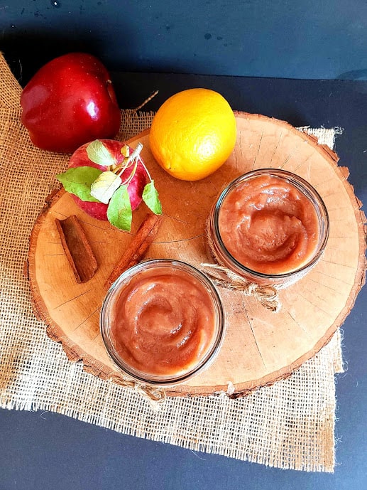 overhead photo shot of homemade apple butter sauce showing caramel colored thick apple butter in two glass container along with few fresh apples and a lemon.