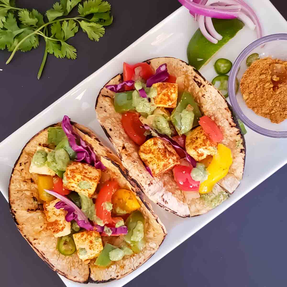 Paneer Fajita Tacos served with green sauce on white plate. This colorful recipe combines Indian Paneer with Mexican Taco seasoning making a lovely fusion.