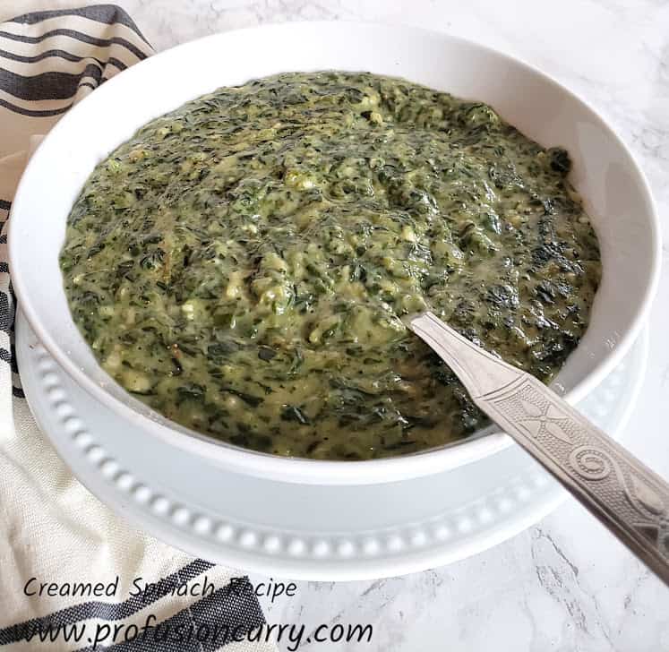 Creamed Spinach served in white serving bowl with serving spoon and kitchen towel on the side.