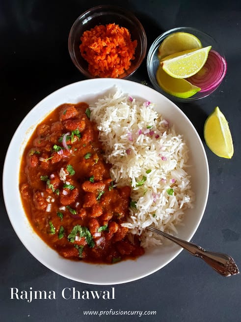 a white serving plate with red beans curry and rice which is garnished with chopped red onions and cilantro leaves. There is carrot pickle and lemon wedges on the side. This comforting Indian meal is called Rajma Chawal or Red Beans and rice.