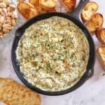 A black cast iron skilet with Cheesy Spinach Dip served as an appetizer tray along with crusty bread slices, crackers and pistachoes. This party spread is perfect for hosting and entertaining.
