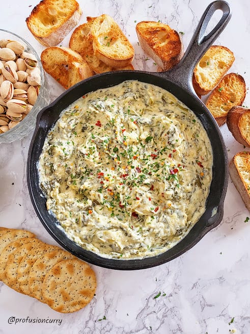 A black cast iron skilet with Cheese Spinach Dip served as an appetizer tray along with crusty bread slices, crackers and pistachoes. This party spread is perfect for hosting and entertaining.