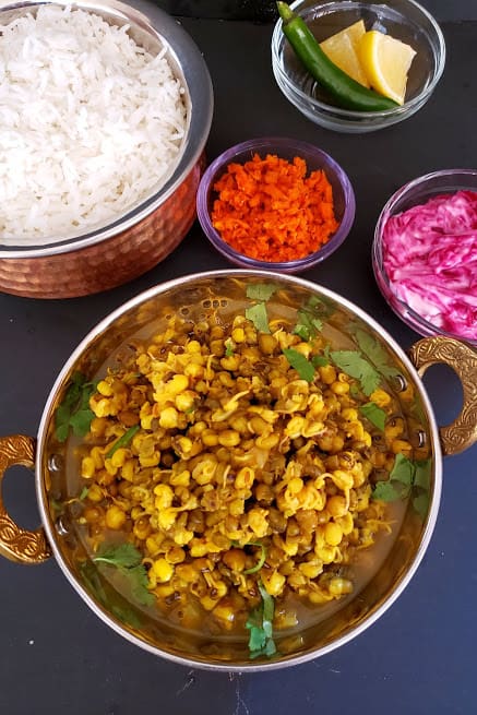 Indian Sprouts Curry served in copper container alog with steamed wihte rice, beetroot raita, carrot pickle and lemon wedges and cut onions. This wholesome meal is classic curry recipe by Profusioncurry.