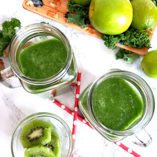 Overhead shot of two glass containers filled with Green smoothie along with pink compostable straws and cut kiti fruit on the side.