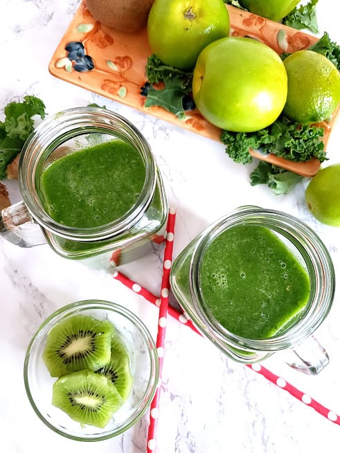 Overhead shot of two glass containers filled with Green smoothie along with pink compostable straws and cut kiti fruit on the side.