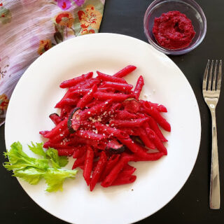 Beet Pesto Pasta served with garnishes on the dinner plate. On the side, there is beet pesto and a fork. A colorful scraf is around the plate.