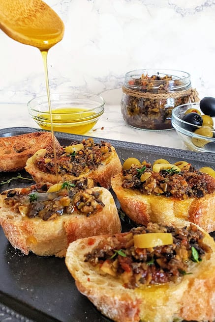 a wooden spoon drizzing olive oil over olive tapenade topped toasts.