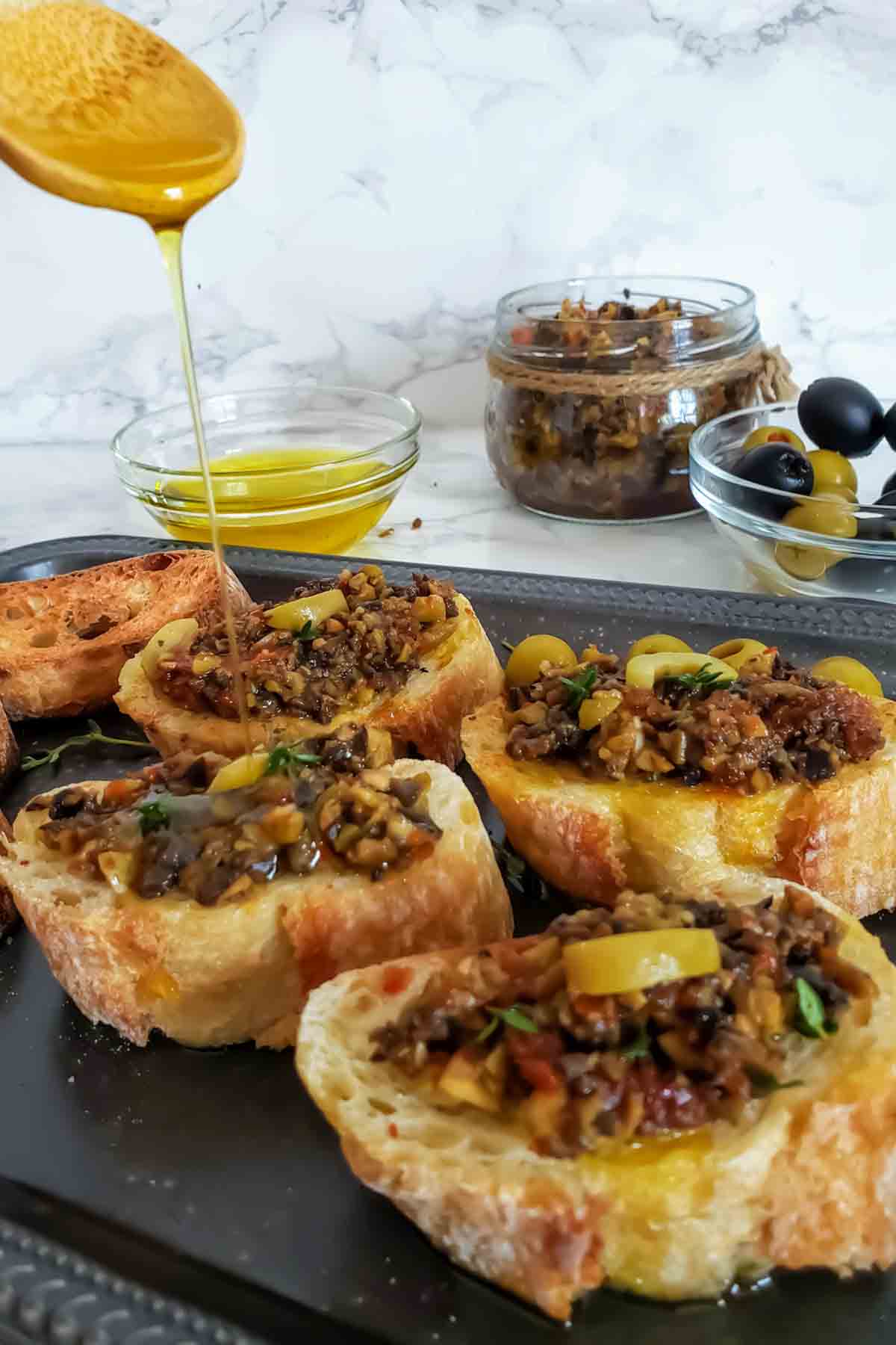 Olive oil getting poured on crostini's topped with vegan olive tapenade.