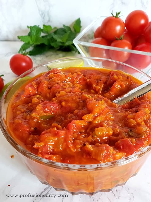 Tomato Chutney served in a glass container with a stainless steel spoon. Fresh mint leaves, cherry tomatoes are in the background.