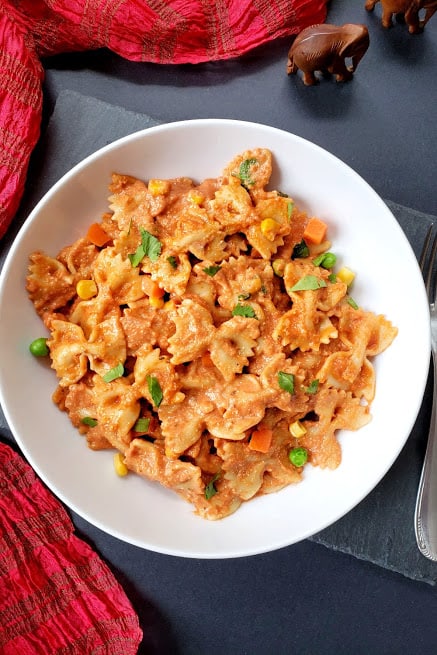 A white dinner plate full of creamy masala pasta dressed up with cilantro leaves and colorful vegetables. A beautiful red scraf is in the background.