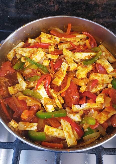 A close up shot of stir fry pan full of colorful veggies and paneer stripes coated in tomato gravy.