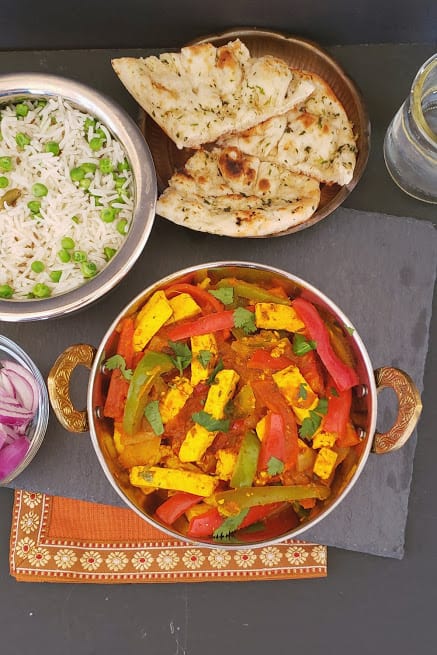 An overhead shot of Indian Dinner including Paneer Jalfrezi in kadhai, basmati rice with peas, naan and cut up onions and lemon.