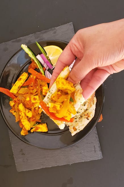 A hand holding a naan and scoop of marinated stir fried paneer on the background of dinner plate of Indian meal.