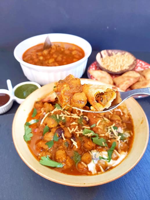 A spoon full of chole, samosa bites and chutneys scooped from the chat serving plate. 