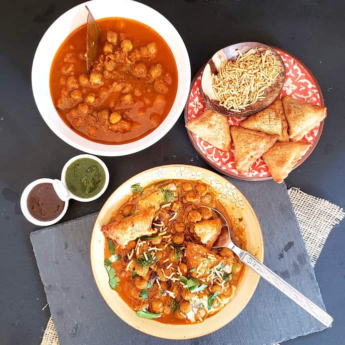 An overhead shot of food serving chickpea curry, baked samosa, two different chuntneys and a prepared plate of layered chat.