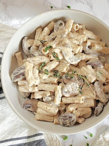 Creamy sauce coated pasta and mushrooms served in white stone bowl with thyme and red chili flakes.