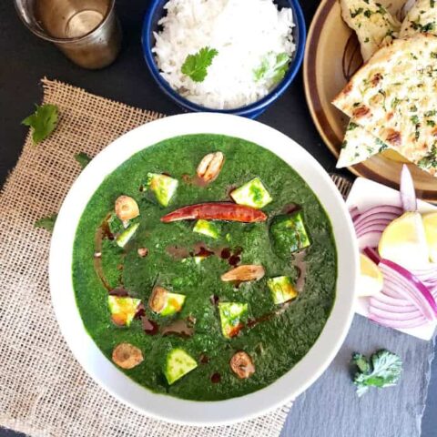 A white bowl full of spinach and paneer curry garnished with oil and garlic tempering. The white steamed rice and garlic naan in the background.