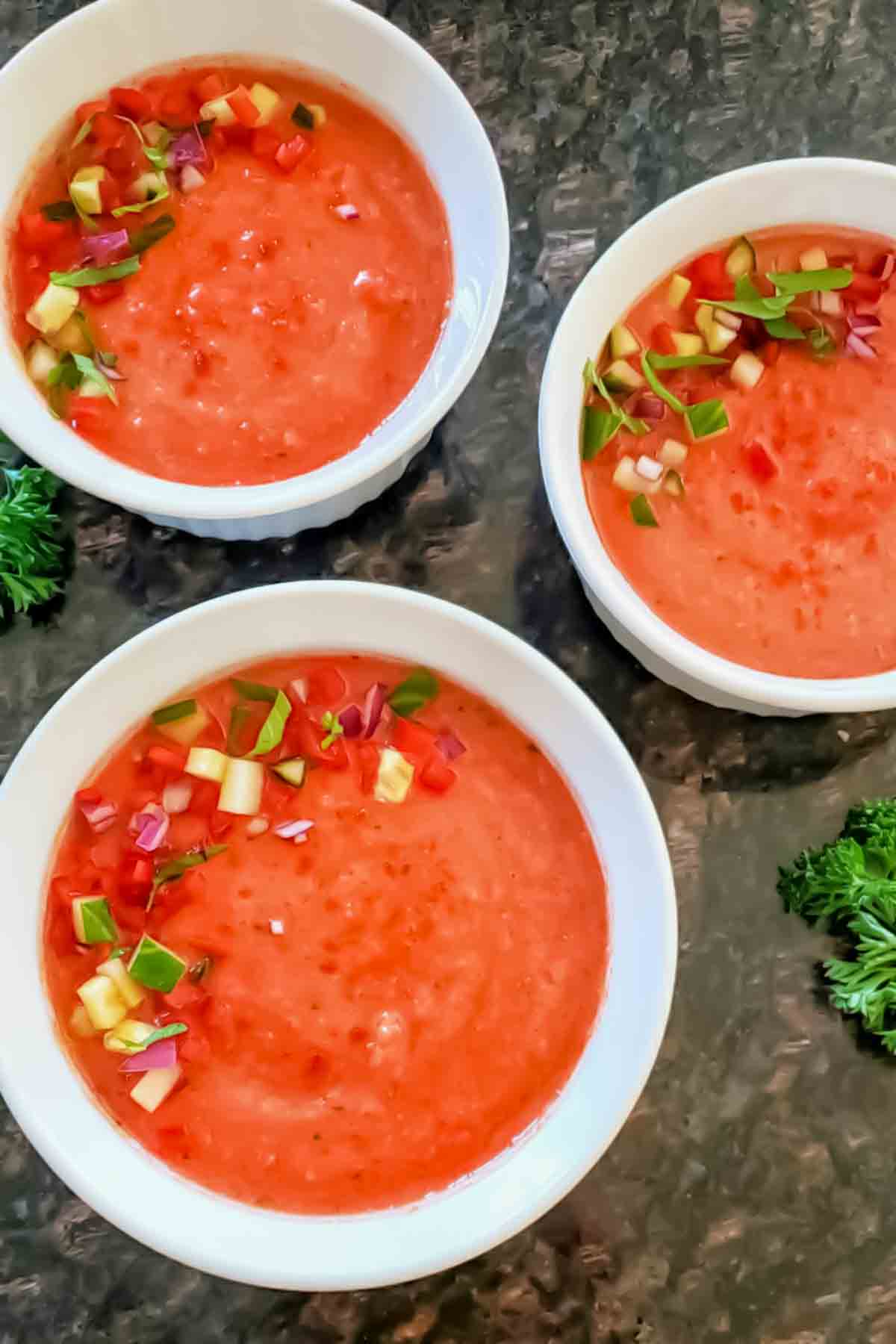 Bowls of vegan gazpacho recipe topped with cucumber, tomatoes, herbs, and olive oil.