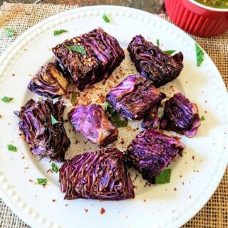 Bunch of air fried red cabbage steaks garnished with red pepper flakes and herbs served on a white dinner plate. A bowl full of chimichurri sauce is in the background for dipping.