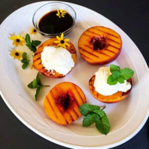 A white serving platter full of grilled peaches, a bowl of balsamic glaze , mint leaves and ice cream.