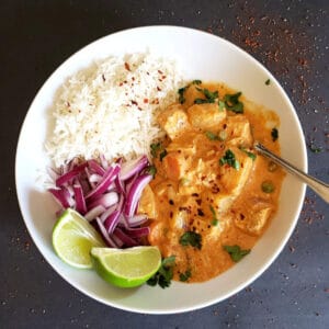 A dinner plate with thai massman curry with vegetables, white rice, red onions and lime wedges.