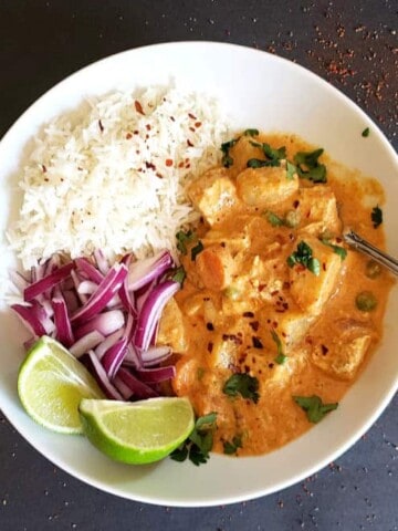 A dinner plate with thai massman curry with vegetables, white rice, red onions and lime wedges.
