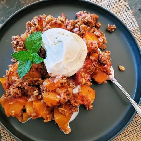 Delicious peach and granola dessert served in a black dessert plate with dessert fork with dollap of vanilla ice cream.