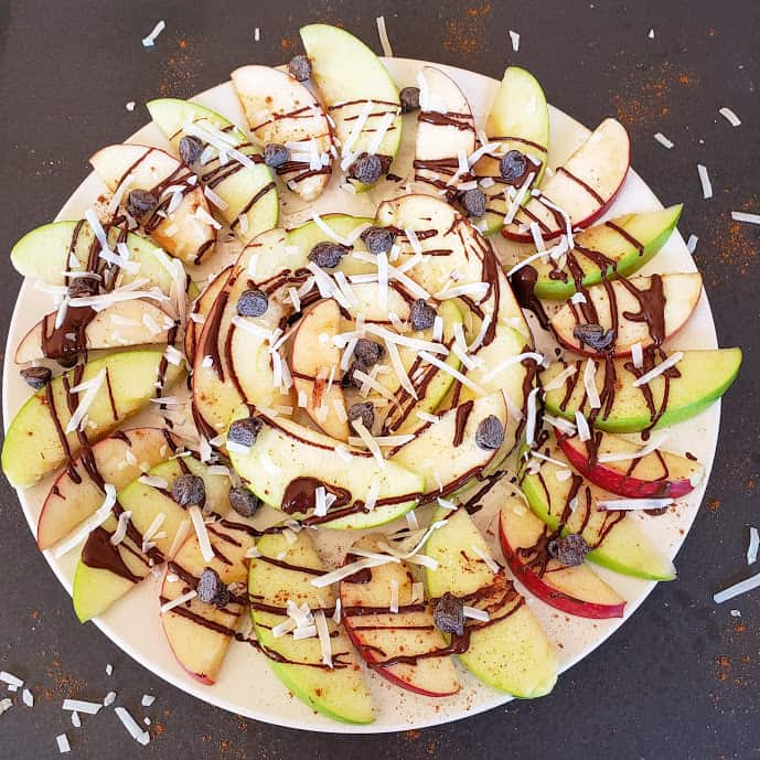 A white platter full of apple slices drizzled with chocolate sauce, chocolate chips, ground cinnamon and coconut flakes. This apple nacho recipe is easy to prepare and tastes amazing.
