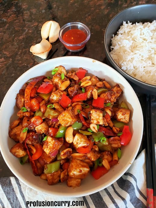 A dinner serving with bowl of kung pao tufu, white rice and red chili sauce along with two fortune cookies.