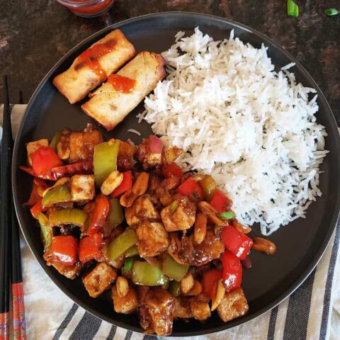 A black round plate with kung pao tofu, white rice and two spring rolls. A chopstick is on the side and dinner napkin under the plate.