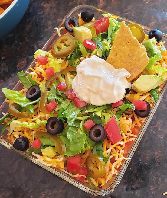 An overhead image of colorful 7 layer dip served in glass container. SHowing the delicious layers of olives, cheese, jalapeno, tomato, lettuce and sour cream with a tortilla chip dipped into this favorite party dip.