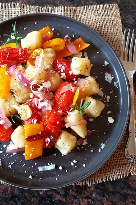 A black dinner plate full of colorful veggies and crisp gnocchi dinner garnsihed with cheese and herbs.