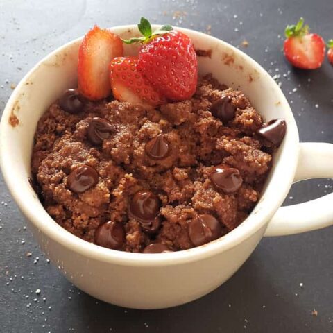 A white mug filled with low carb, vegan almond flour chocolate dough and microwaved to make a mug cake. It is garnished with sliced strawberries to make decadent single serve dessert.
