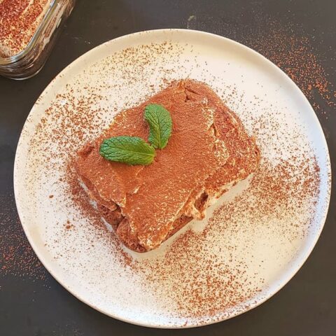 A white dessert plate with a slice of layered tiramisu dessert served with mint leaves and cocoa powder dusting on the top.