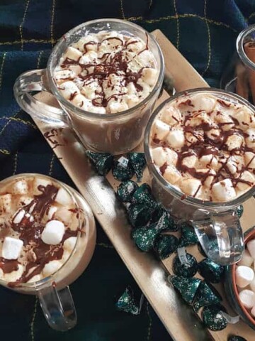 Homemade hot chocolate drink served in three glass mugs along with chocolate bites and marshmallows served on silver tray. The whole set up is served on cozy blanket.