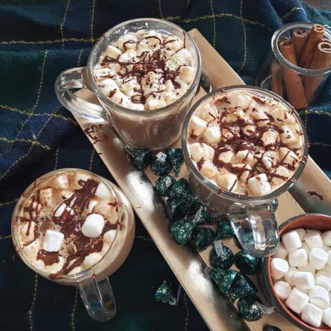 Homemade hot chocolate drink served in three glass mugs along with chocolate bites and marshmallows served on silver tray. The whole set up is served on cozy blanket.