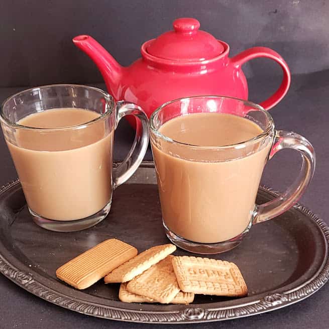 A red tea kettle and two glass cups filled with Indian Spiced tea latte. This masala chai is served with tea cookies.