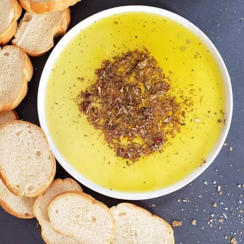 Italian Bread Dipping oil with herbs, garlic and vinegar served on white plate with crusty bread pieces on the side.